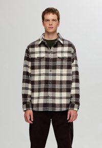 Flannel shirt in cream and dark brown plaid, featuring a button-up front, collar, and two chest pockets. Soft, textured fabric.