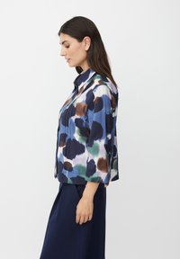 Donna con capelli lunghi e scuri che indossa una camicia button-up a fantasia a macchie blu, verde, marrone e bianca e pantaloni blu navy, in piedi di profilo.