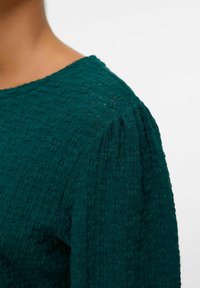 Tyg med textur i teal med rund halsringning och lätt puffärmar. Tyget har ett vävt mönster som ger djup och dimension.