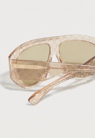 Lunettes de soleil surdimensionnées beige transparent avec des verres marron clair et un motif de logo répétitif en relief sur la monture et les branches.
