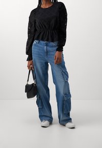 Schwarze strukturierte Bluse mit puffärmel, kombiniert mit hellblauen Cargo-Jeans mit mehreren Taschen. Ergänzt durch eine kleine schwarze Handtasche.