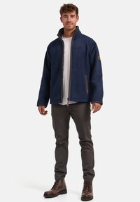 Marineblaue Fleecejacke mit hohem Kragen, Reißverschluss und braunen Lederakzenten. Bietet eine lockere Passform und zwei seitliche Taschen.