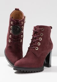 Botas de tobillo de ante burdeos con puntera redondeada, ojales dorados, cordones en la parte delantera y tacón de plataforma negro. Presenta un pompón decorativo negro.