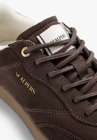 Bruna mockaskor med guld "SCALPERS"-logga, kontrasterande beige gummisula och släta bruna snören. Inredningen har vit textillining.