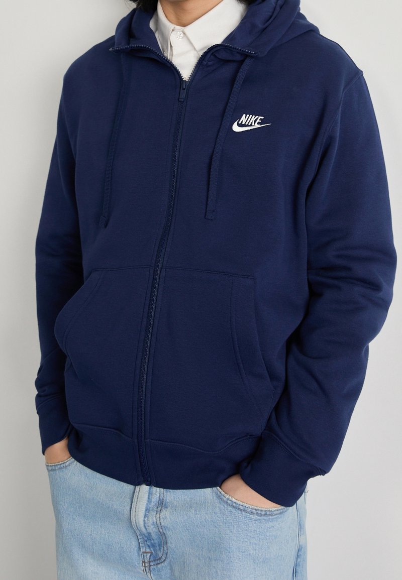 Námořnická modrá mikina s kapucí na zip vyrobená z měkkého materiálu, s klokankovou kapsou a bílým logem Nike na horní levé straně.