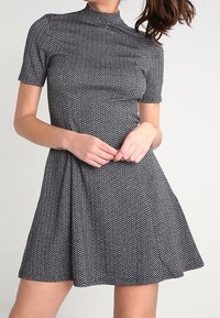 Femme portant une robe en maille à manches courtes, motif zigzag noir et blanc, col montant et jupe évasée.