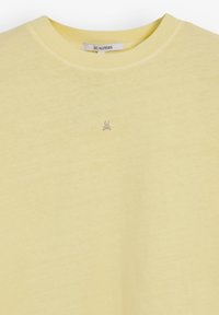 Žluté tričko vyrobené z měkké, lehké tkaniny s kulatým výstřihem. Na hrudi je malé logo lebky v kontrastní barvě.