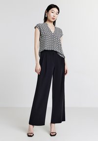Donna che indossa pantaloni neri a gamba larga, una blusa a maniche corte bianca e nera con motivo a diamante e tacchi aperti neri, in piedi contro uno sfondo semplice.