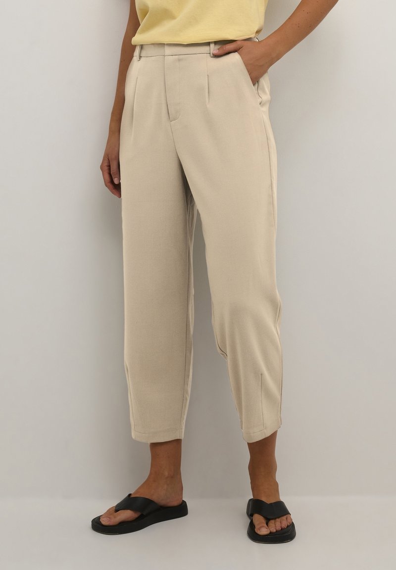 Femme portant un pantalon beige plissé taille haute et longueur cheville avec des sandales noires à enfiler, debout devant un fond gris clair.