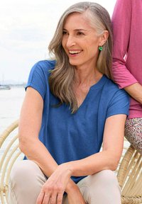 Blauwe V-hals T-shirt met korte mouwen, gemaakt van een gladde stof. Model heeft lang grijs haar en zit naast een persoon in een roze shirt.