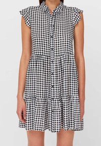 Schwarz-weißes Gingham-Hemdblusenkleid mit kurzen Rüschärmeln und gestuftem Rock, getragen von einer Person mit hängen gelassenen Armen.