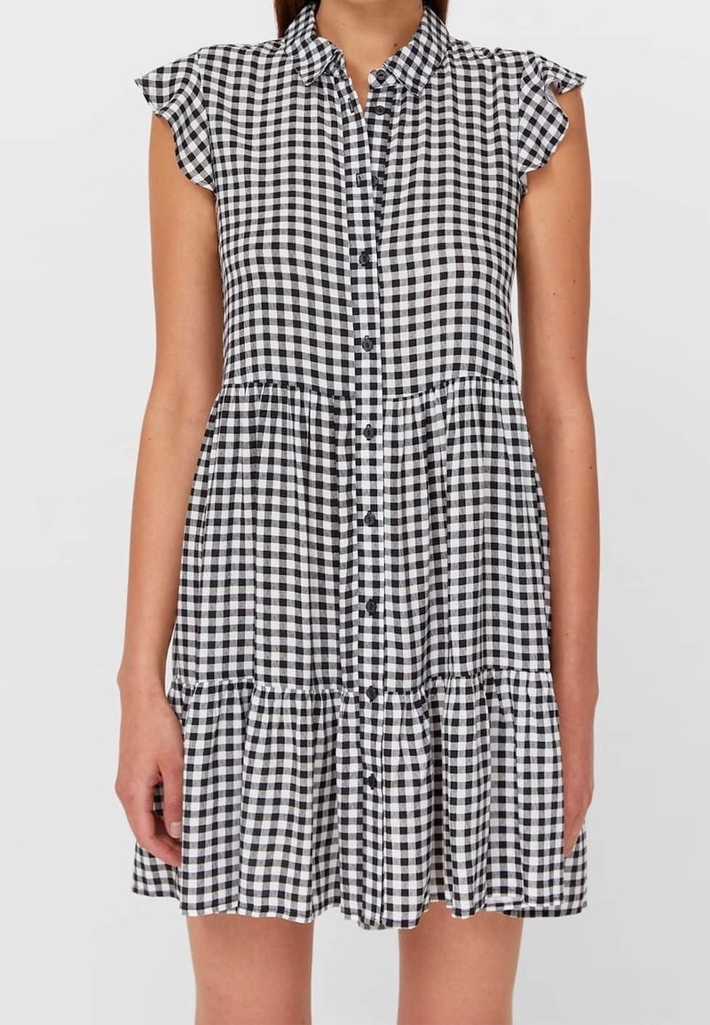 Schwarz-weißes Gingham-Hemdblusenkleid mit kurzen Rüschärmeln und gestuftem Rock, getragen von einer Person mit hängen gelassenen Armen.