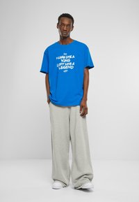 Blå grafisk t-shirt med fronttekst i hvid, der siger "JEG SPILLEDE SOM EN KONGE, FORLADTE SOM EN LEGEND"; parret med lysegrå baggy bukser.