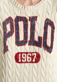 Gros plan sur un pull crème en maille torsadée avec "POLO" en grandes lettres bleu marine et rouges, et "1967" en rouge en dessous sur la poitrine.