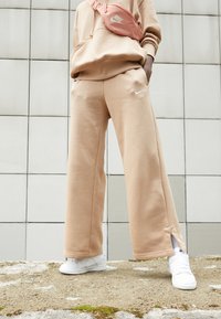Pantalones de pierna ancha en beige claro con un logo blanco de Nike, combinados con zapatillas blancas y una riñonera rosa a juego, sobre una superficie texturizada al aire libre.