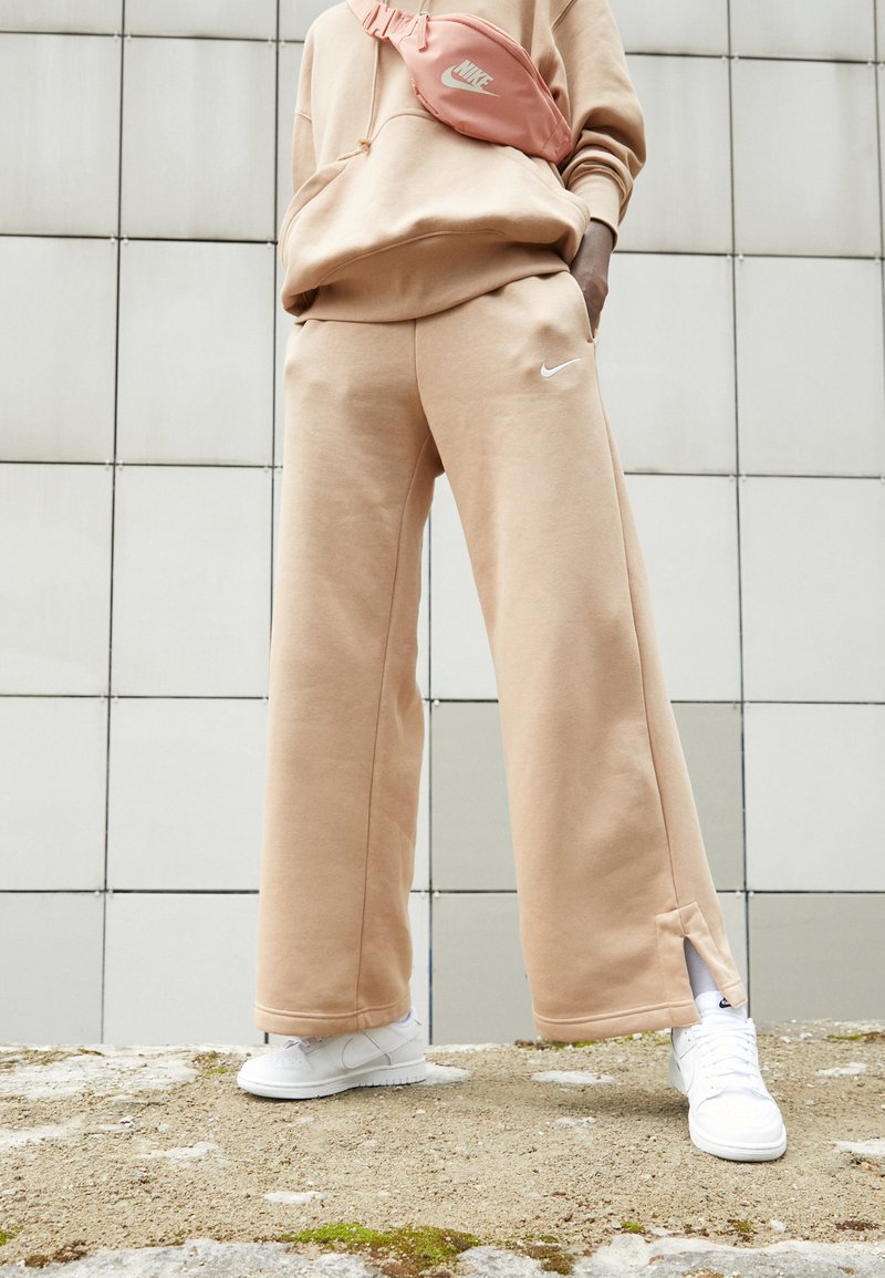 Pantalones de pierna ancha en beige claro con un logo blanco de Nike, combinados con zapatillas blancas y una riñonera rosa a juego, sobre una superficie texturizada al aire libre.