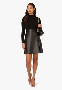 Schwarzer Kunstledrock mit tailliertem Bund und weit auslaufendem Saum. Kombiniert mit einem langärmligen schwarzen Oberteil und nudefarbenen Absatzsandalen. Kleine schwarze Handtasche.