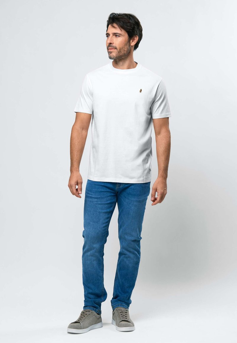 Homme debout, portant un t-shirt blanc uni, un jean bleu et des baskets grises, regardant légèrement sur le côté sur un fond uni.