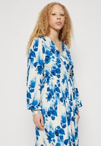 Blomsterkjole med hvit bakgrunn og store blå blomster, lange ermer med rynkede mansjetter og omslagsutringning.