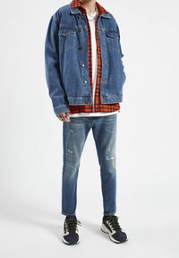 Chaqueta de mezclilla, azul con parches desgastados; debajo, una camisa de cuadros en rojo y negro en capas. Jeans azules descoloridos y zapatillas negras/azul marino con detalles.