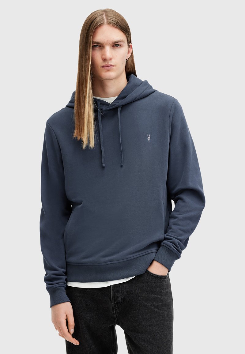 Navy blue hoodie made of soft fabric, featuring a kangaroo pocket, drawstrings, and a small logo on the chest. Worn with black jeans.