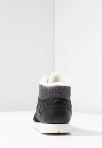 Svart och grå quiltad hög sneakers med vit pälsfoder och vit sula, visad bakifrån på en vit yta och bakgrund.