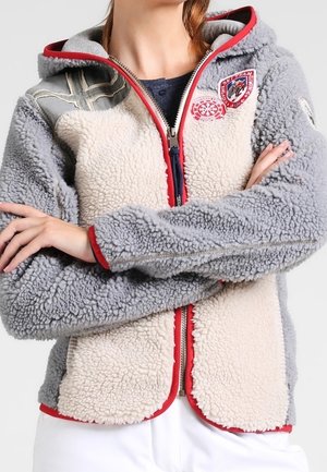 Person iført en grå og beige fleecejakke med røde kant og patches, armene krydset, og hvide bukser synlige nederst.