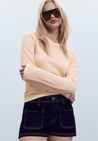 Svetlo oranžna majica z dolgimi rokavi iz mehkega materiala; temnonavaje modri šortsi z rumenim šivanjem; model s svetlimi lasmi in velikimi sončnimi očali.