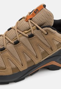 Tan mesh and leather hiking shoe with black accents. Features a textured upper, orange pull tab, and rubber outsole for traction.