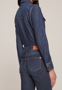 Denim jumpsuit in dark blue with long sleeves, back details, and a leather patch on the waistband. Features classic stitching and button accents.