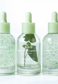 Drei klare Tropfflaschen mit der Aufschrift Abib Heartleaf TECA Kapselserum und grünen Verschlüssen; in der mittleren Flasche befindet sich ein grünes Pflanzenblatt.