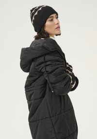 Manteau rembourré noir avec un design matelassé, doté d'une capuche et d'une fermeture éclair. Le modèle porte un bonnet en tricot à motifs noir et beige.