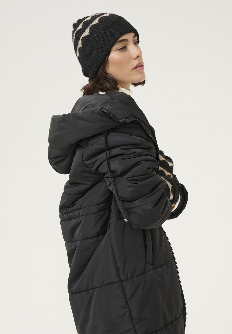 Manteau rembourré noir avec un design matelassé, doté d'une capuche et d'une fermeture éclair. Le modèle porte un bonnet en tricot à motifs noir et beige.
