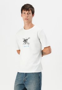 Hvid T-shirt med korte ærmer og grafisk sort skull iført en hat og krydsede knogler. Har blå tekst nedenunder. Enkel, afslappet design.