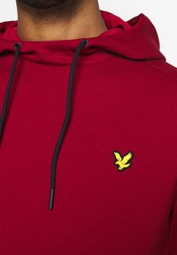 Roter Hoodie mit schwarzen Kordeln, ausgestattet mit einem gelben gestickten Vogel-Logo auf der linken Brust. Der Stoff sieht weich und strukturiert aus.