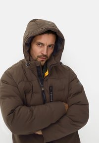 Brun isoleret dunjakke med hætte, forsynet med lynlås foran, sidelommer og kontrasterende gul inderforing. Struktureret stof.