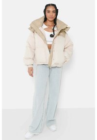 Giacca a vento di colore beige chiaro con un ampio colletto, zip frontale e bottoni a pressione. Indossata sopra un crop top bianco, abbinata a jeans azzurri.