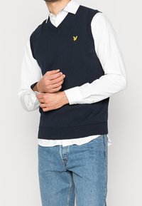 Suéter sin mangas de punto azul marino con diseño de cuello en V y dobladillo acanalado. Presenta un pequeño logo de un pájaro amarillo en el pecho izquierdo. Se lleva sobre una camisa blanca.