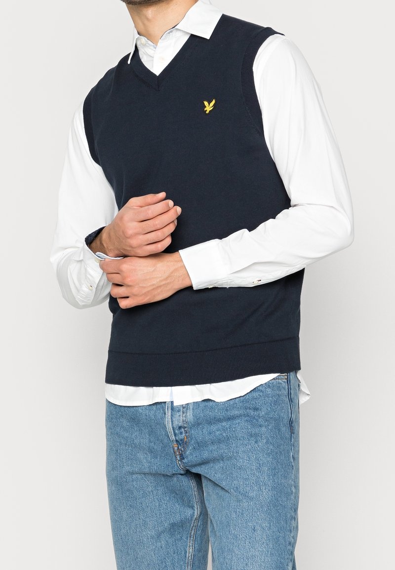 Suéter sin mangas de punto azul marino con diseño de cuello en V y dobladillo acanalado. Presenta un pequeño logo de un pájaro amarillo en el pecho izquierdo. Se lleva sobre una camisa blanca.