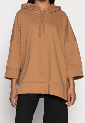 Mujer con sudadera holgada de color camel con mangas tres cuartos y pantalones negros sobre fondo liso.