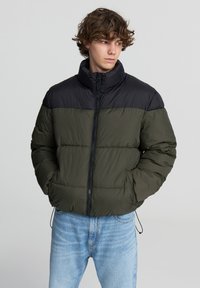 Giacca a vento con sezione superiore nera e parte inferiore verde oliva, colletto alto, zip frontale e tasche laterali. Design trapuntato e testurizzato.