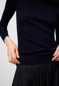 Granatowy sweter dzianinowy z długimi rękawami, dopasowany krój i miękka tekstura. Połączony z plisowaną czarną spódnicą w talii, ręce delikatnie dotykają materiału.