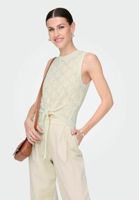 Top tejido de color verde claro con un detalle de lazo en la cintura, presenta un diseño sin mangas y un patrón texturizado. Combinado con pantalones beige y un bolso marrón.