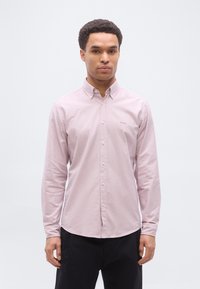 Άνδρας που φοράει ένα ανοιχτό ροζ πουκάμισο με κουμπιά και μακριά μανίκια και μαύρο παντελόνι, στέκεται μπροστά σε μια απλή γκρι ανοιχτή φόντο.