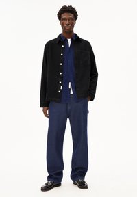 Zwarte corduroy knoopjack over een blauwe shirt, gecombineerd met blauwe spijkerbroek en zwarte schoenen. Opvallende texturen en kleuren zijn aanwezig.