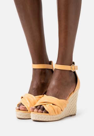 Sandales compensées en cuir jaune avec un design noué à l'avant, base en espadrille tissée et fermeture à lanière autour de la cheville.