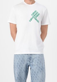 T-shirt en coton blanc avec un logo diagonal vert affichant le mot "calvin" répété. Encolure ronde, manches courtes, coupe décontractée.