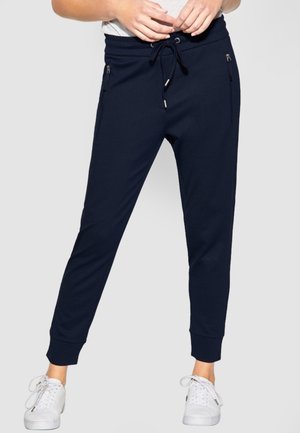 Personne portant un pantalon de jogging bleu marine avec taille à cordon et poches latérales zippées, porté avec des baskets blanches, debout devant un fond uni.