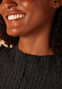 Blouse en tissu noir texturé avec un design froncé autour de l'encolure. Gros plan montrant un visage souriant avec des cheveux bouclés texturés et une boucle d'oreille.