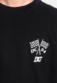 Black cotton t-shirt featuring white graphic design of crossed checkered flags and "DC 94" text, with a DC logo underneath.